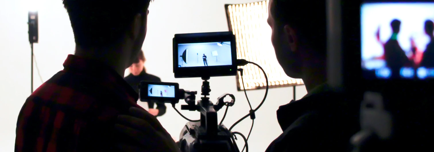 Two people filming a person in a studio with lights and cameras showing the scene on monitors.