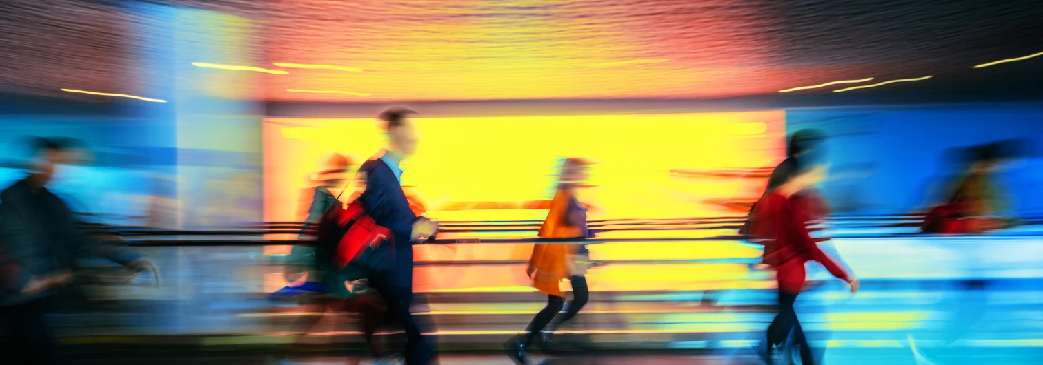 Blurred image of people walking quickly in a brightly lit indoor urban area with colorful lights.