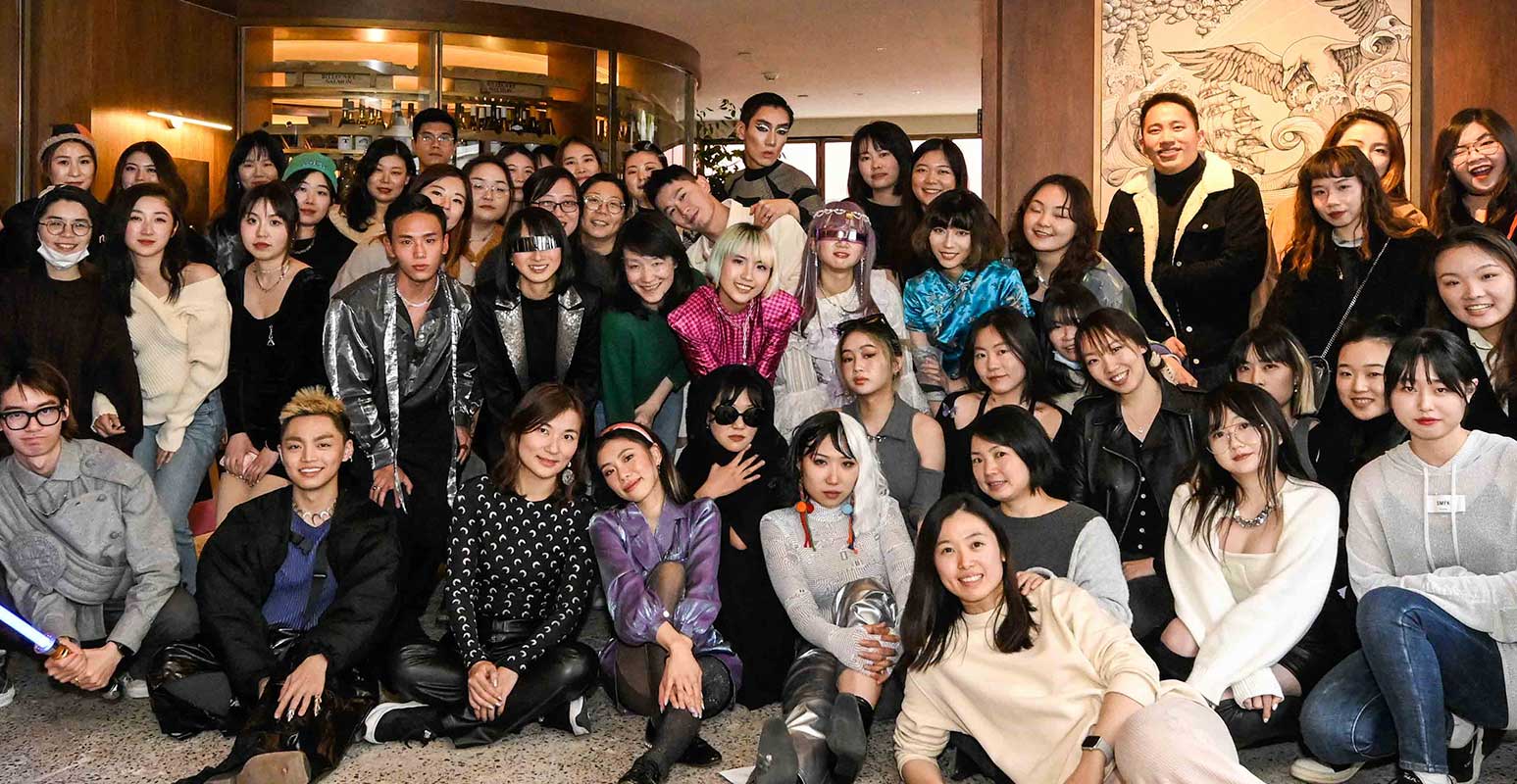 Large group of diverse young adults posing indoors with smiles, some wearing distinctive fashion and accessories.